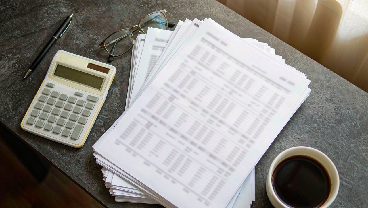 Printed financial statements in two stacks on a desk with a calculator, pen, and cup of coffee in window light