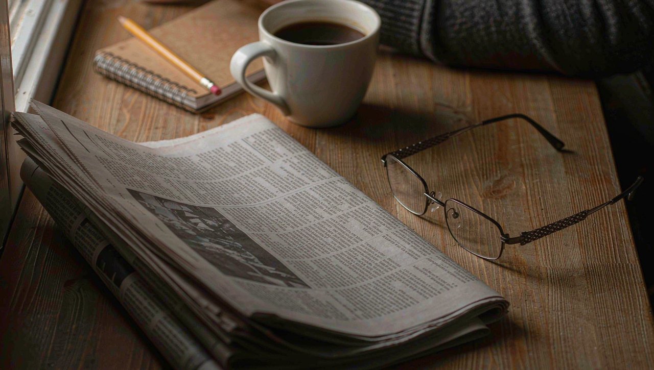 A financial newspaper market section on a desk beside reading glasses and a cup of coffee