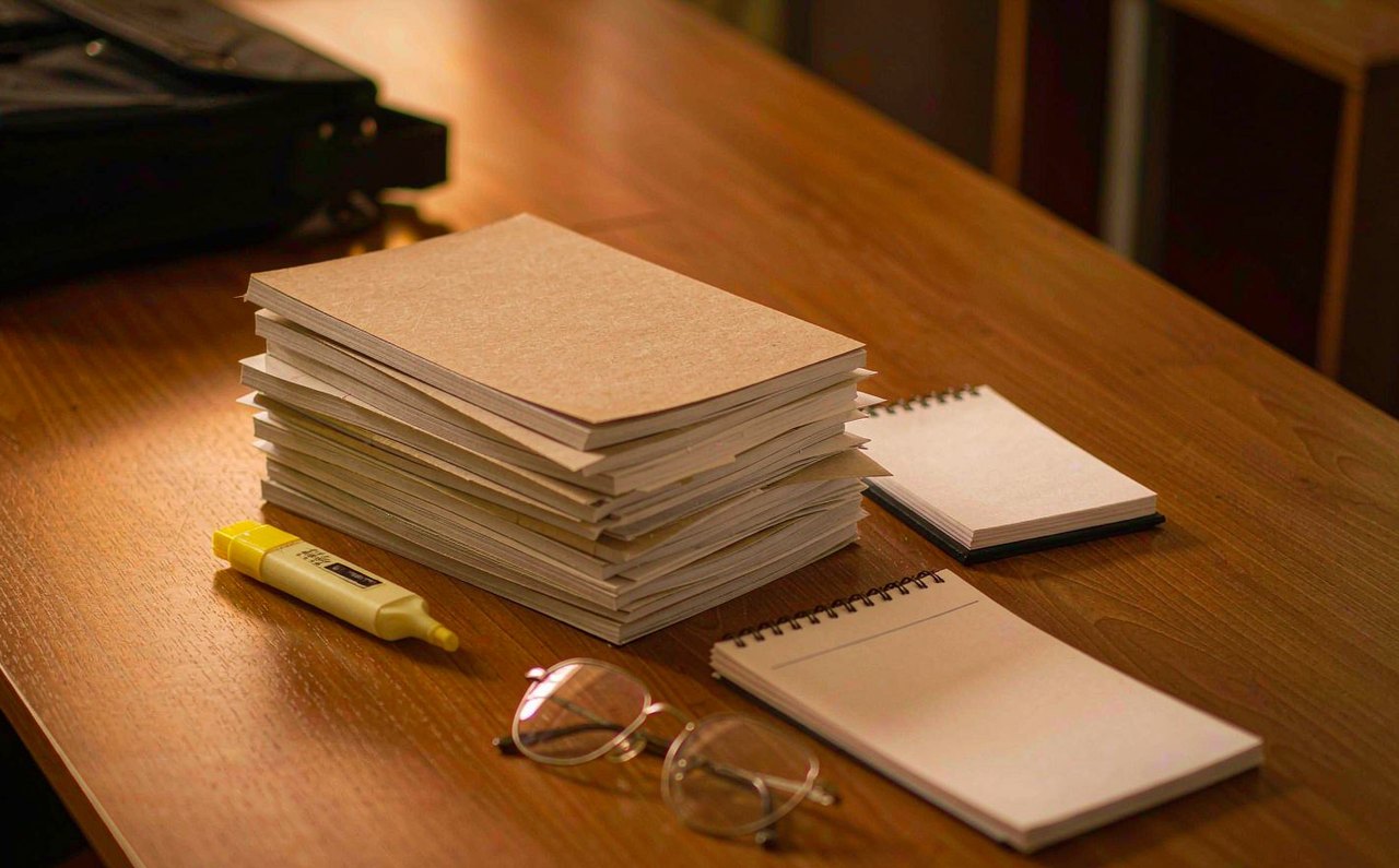 A thick stack of plain annual report booklets on a walnut desk in warm lamp light with a highlighter and reading glasses