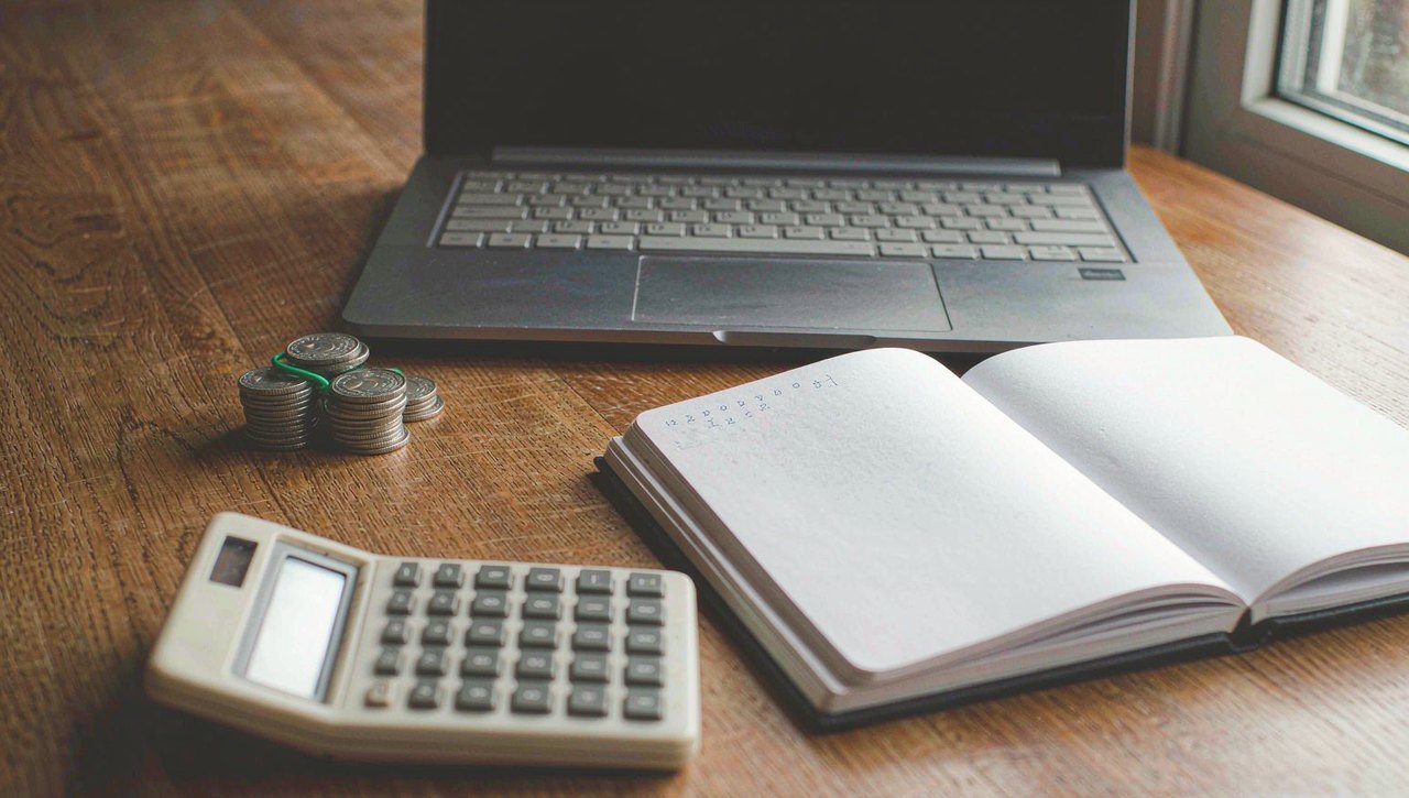 A calculator, a notebook with handwritten percentages, and a few coins on a wooden desk beside a laptop
