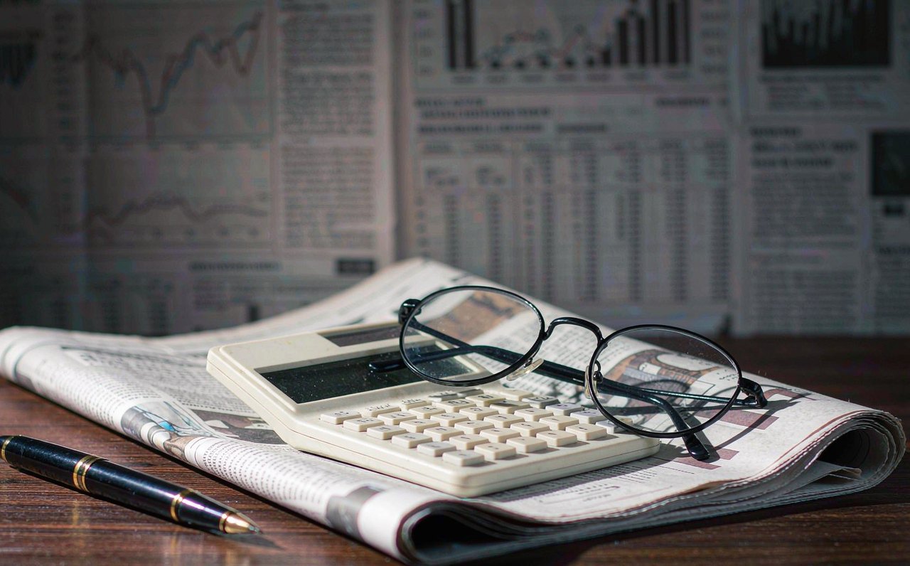 A financial newspaper-style layout of blurred stock charts on a desk beside a calculator and reading glasses