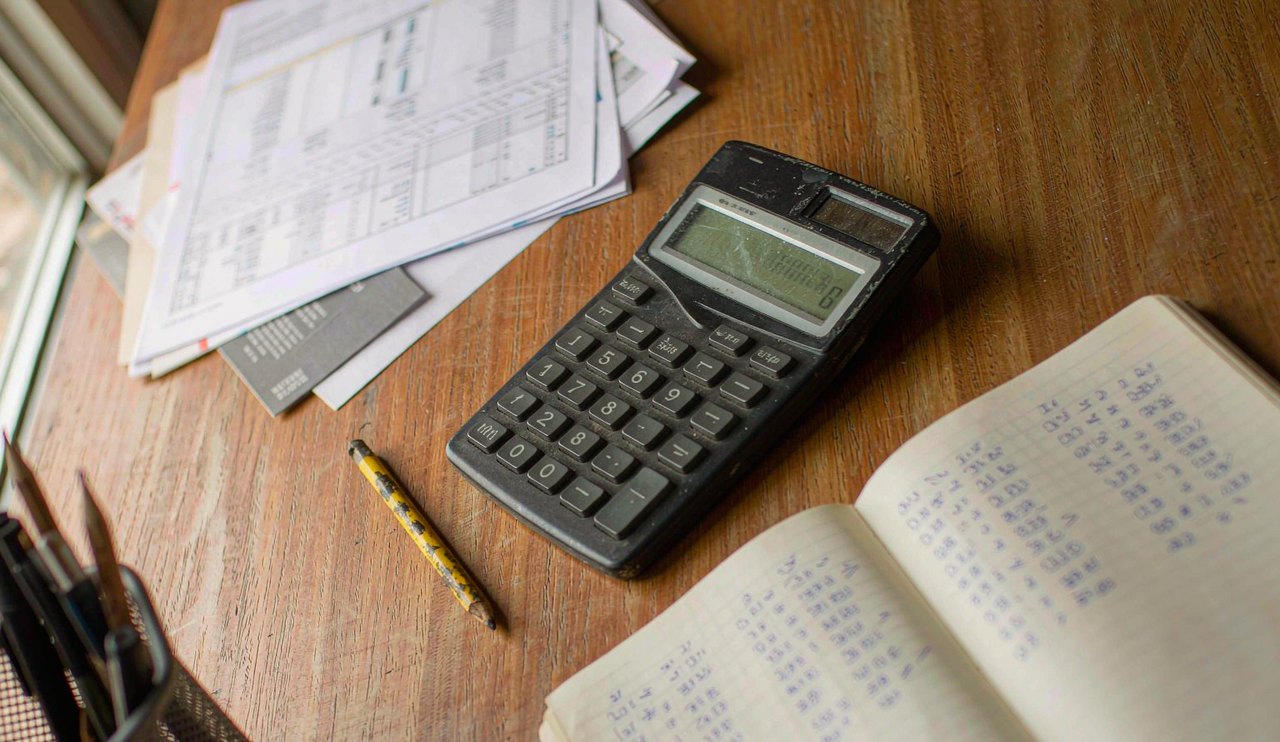 Printed account statements, a calculator and a notebook with two columns of figures on a wooden desk
